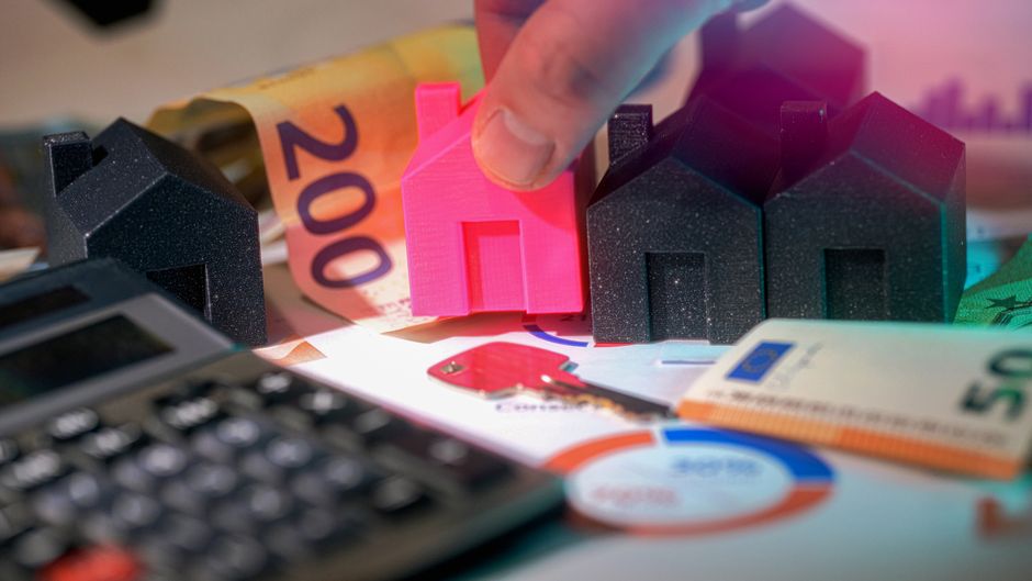 A close-up view of a person's hand placing a small pink model house on a cluttered surface that includes three larger model houses, one black and two dark purple, all made of textured, matte material.