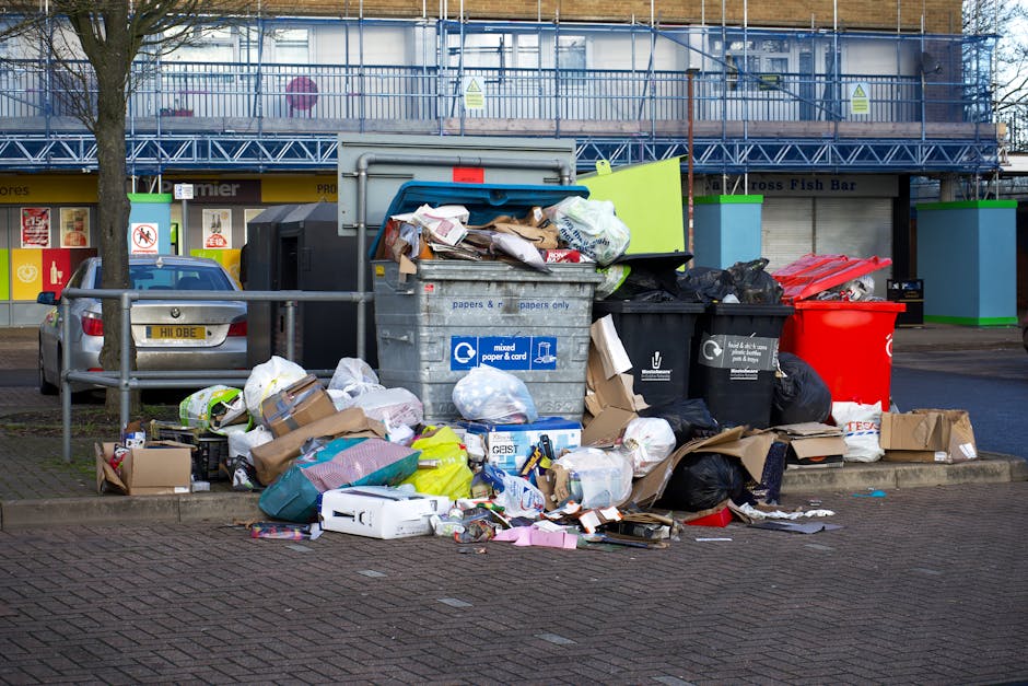 A large outdoor pile of mixed household waste consisting of discarded plastic containers, bottles, and tubs in various colors such as white, blue, pink, green, and red, scattered across a paved ground
