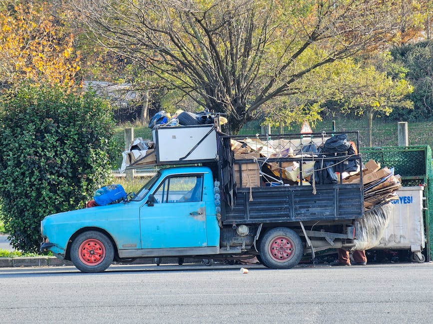 A small, old blue pick-up truck with faded and peeling paint sits parked on the side of a street, loaded with a large, overflowing collection of discarded household items and miscellaneous rubbish. Th