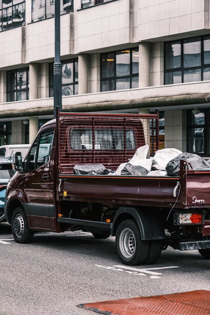 A small, old blue pick-up truck with faded and peeling paint sits parked on the side of a street, loaded with a large, overflowing collection of discarded household items and miscellaneous rubbish. Th