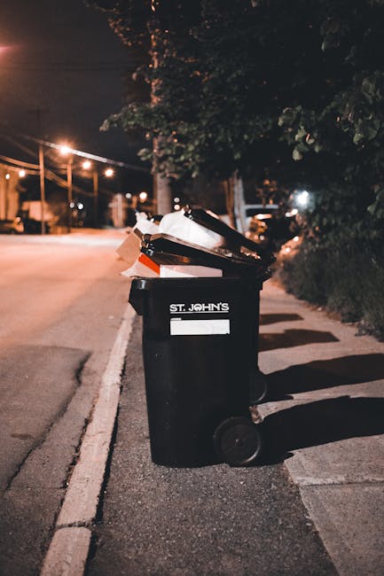 A black outdoor rubbish bin with the label 'St. John's' positioned on a sidewalk at night, slightly to the right of the scene. The bin is filled with a mix of household waste, including cardboard boxe